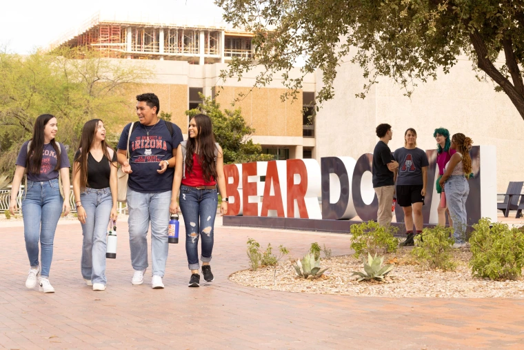 Group of University of Arizona students walking and talking on campus in front of large red and white “Bear Down” letters, with another group standing and chatting nearby.